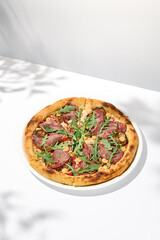 Italian pizza with roast beef, arugula and spicy sauce on light background with shadows. Meat pizza with sliced roast beef on white table. Italian food in trendy menu restaurant