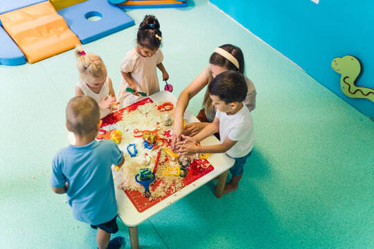 Calming Sensory Play With Moldable Kinetic Sand At Nursery School. Toddlers With Their Teacher Having Fun Around The Table Using Different Tools For Sculpting Sand Such As Colorful And Textured