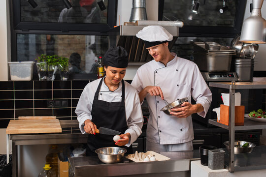 african american sous chef holding knife near raw egg above bowl while cooking near colleague in kitchen. - Powered by Adobe