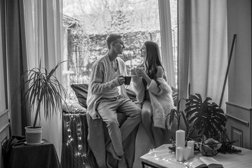 A young and attractive couple, a guy and a girl, are sitting on the windowsill in blankets and warm sweaters in winter during a snowfall, hugging and drinking tea. Emotions and feelings