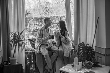A young and attractive couple, a guy and a girl, are sitting on the windowsill in blankets and warm sweaters in winter during a snowfall, hugging and drinking tea. Emotions and feelings