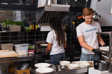 Fototapeta na wymiar happy redhead volunteer cooking near african american woman in kitchen.