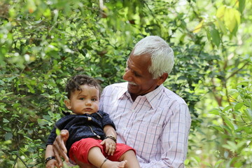 Naklejka na ściany i meble Picture of a proud grandfather holding his Adorable grandson and looking at him with love. Standing in a backyard of green a farm.