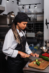 side view of african american woman in apron cutting cucumber in modern kitchen.
