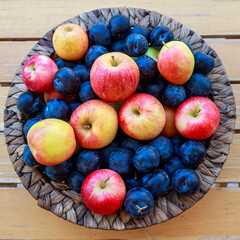 Big plate of of ripe blue plums and red yellow apples on wooden table, top view
