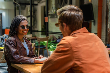 happy african american woman in sunglasses looking at blurred friend in outdoor cafe terrace.
