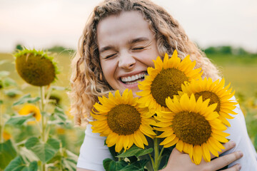 Young caucasian woman with curly hair posing while holding some sunflowers in a blooming field of...