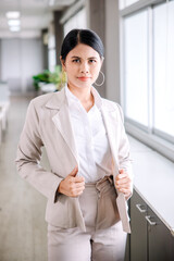 A young businesswoman stands and looks at the camera with a confident expression in the office.