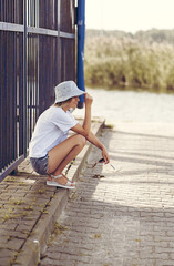 Attractive woman in a white t-shirt and bucket hat sits in the shade on the bank of the river, smiling, looking at the water. for a serene and relaxing outdoor concept