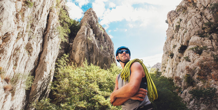 Portrait of smiling climber man in protective helmet and sunglasses with climbing rope on the shoulder standing in Paklenica park between rock cliff walls. Active extreme sports time spending concept.