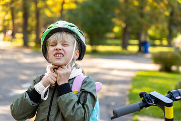 Profile view portrait cute blond little caucasian school girl putting on wrong size painful helmet riding electric scooter city street park outdoors sunny day. Healthy sport children activity outside