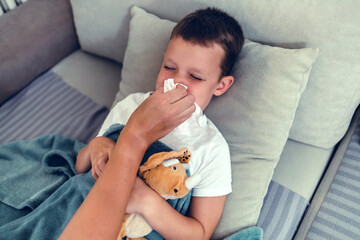 Sick, ill and unwell little boy suffering from cold, flu or covid and lying on the sofa at home while blowing his nose with mom. Cute son with a runny nose and resting in the living room from above.