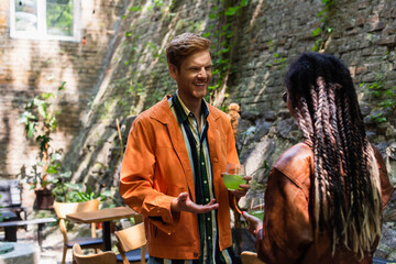 Obraz premium happy redhead man looking at african american woman while gesturing and holding glass with green cocktail outside.