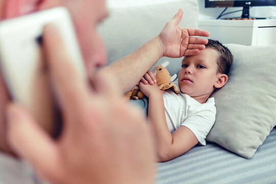 Worried man talking on smartphone while sitting near diseased son in the dedroom. Worried father consulting doctor on cellphone while sitting next to his little son in the living room.