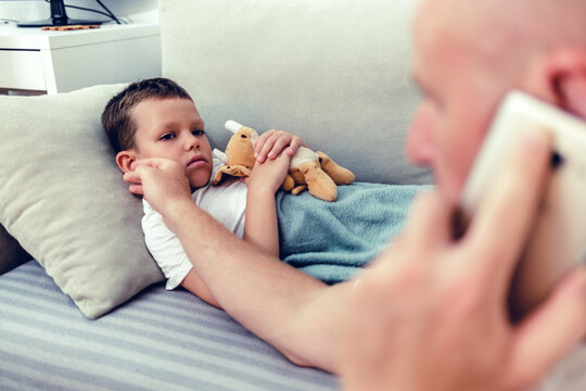 Worried man talking on smartphone while sitting near diseased son in the dedroom. Worried father consulting doctor on cellphone while sitting next to his little son in the living room.