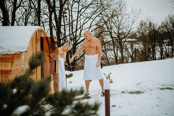 Senior woman in towel with her husband coming out from outdoor sauna during cold winter day. - obrazy, fototapety, plakaty