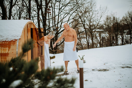 Senior woman in towel with her husband coming out from outdoor sauna during cold winter day.