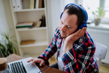 Young man with Down syndrome sitting at desk in office and using laptop, listening to music from...