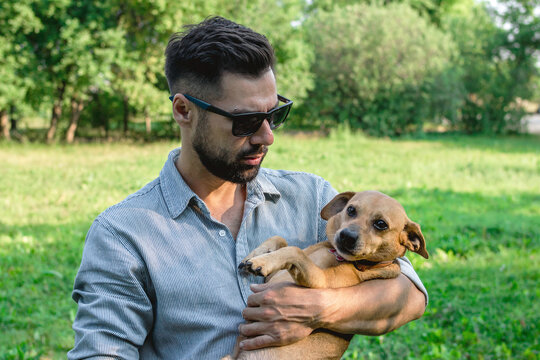 Handsome stylish European man is holding his dog in hands in park on a walk. Friendship between human and pets.