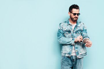 Portrait of handsome confident stylish hipster lambersexual model. Sexy man dressed in jeans jacket. Fashion male isolated on blue wall in studio. In sunglasses