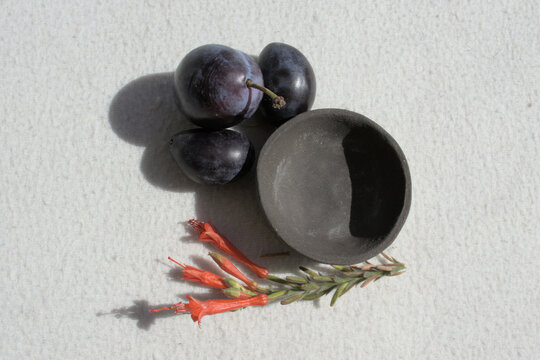 Three Plums, A Cup And Small Orange Flowers Seen From Above.