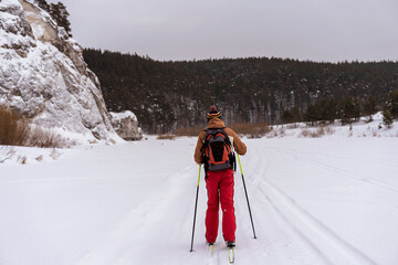 Rear view of young man in red brown clothes with backpack skiing near rocks and cliffs Active healthy lifestyle Winter sports Hiking