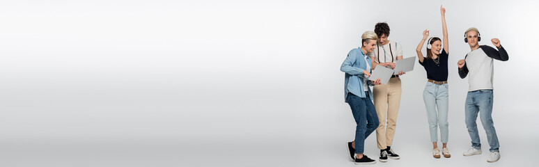 cheerful couple dancing in wireless headphones near friends with laptops on grey background, banner.