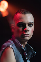 Close-up portrait of young man wearing makeup, smokey eyes, posing isolated over dark background in neon. Spot light