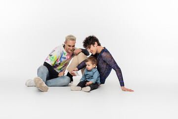 Fototapeta na wymiar happy gay men sitting with toddler kid and taking selfie on smartphone on grey background.