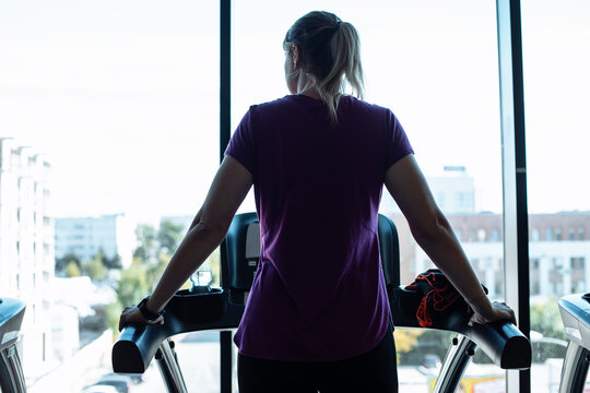 Plus size woman alone run on treadmill at fitness gym and look out window at city life, back view. Overweight girl do cardio workout on training apparatus. Weight loss, fat burning, sport lifestyle - Powered by Adobe