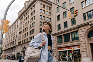 Fototapeta na wymiar Stylish modern caucasian woman in blue shirt with backpack walking in european city. Positive girl spends time on city street. Brunette looks to side, wears casual clothes. Student concept