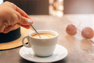 Cappuccino on the table in cafe. Women's hands stirs the sugar in coffee. Morning sunlight falls on table. Breakfast concept. Selective focus