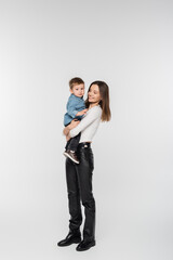 full length of young and happy woman holding toddler son on grey background.