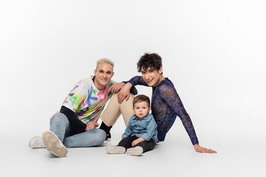 stylish gay partners sitting near toddler son on grey background.