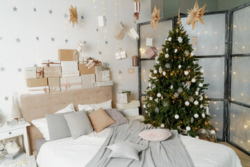 Pile of Christmas gifts in colorful wrapping with ribbons against a white wall with silver stars. Christmas tree in the room.