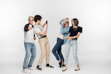 Fototapeta premium young men taking photo on smartphones near cheerful women dancing on grey background.