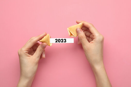 Woman holding tasty fortune cookie and paper with number 2023 on pink background