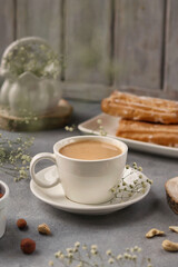 A white cup of coffee with milk stands on a gray background, behind a plate with eclairs