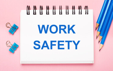 On a light pink background, light blue pencils, paper clips and a white notebook with the text WORK SAFETY