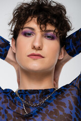 portrait of brunette queer person with makeup and hands behind head isolated on grey.