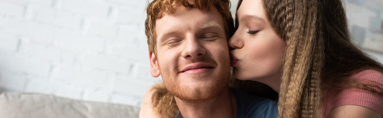 pretty teen girl hugging and kissing cheek of redhead boyfriend in living room, banner.