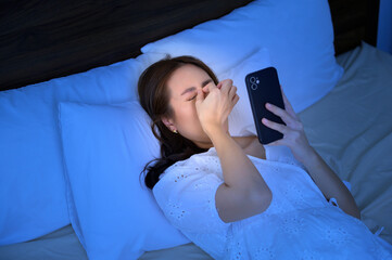 asian woman feeling eye strain fatigue while using smartphone in bedroom