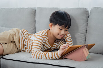 Handsome Asian boy, lying on sofa in his comfortable position, along with hands on feet, chin, Boy laying down watching online clips on his tablet merrily, his face looked happy.