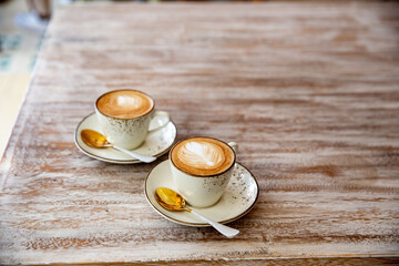 Two stylish white cups of cappuccino with latte art on saucer and golden spoons on light wooden...