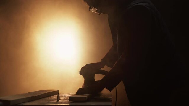Woodwork hobby. Carpentry production. Retirement hobby. Senior joiner in goggles polishing wood using sander tool in dark workshop with glowing smoke cloud.