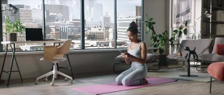Young African American sporty young woman sits on floor in living room, using yoga online fitness training application to get the workout plan