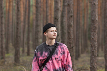 Thoughtful young man in casual clothing and black hat with shoulder belt bag walking in a pine autumn forest.
