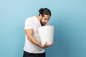 Portrait of man with beard wearing white T-shirt vomiting. holding bucket in hands, suffering...