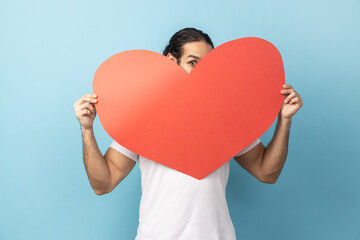 Portrait of man with beard wearing white T-shirt peeking out of big read heart with flirting look and expressing love, greeting on Valentine's day. Indoor studio shot isolated on blue background.