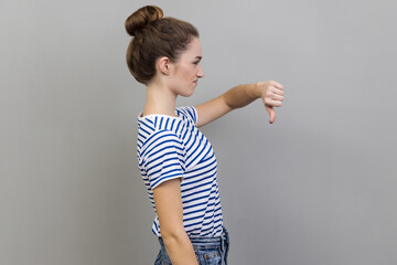 Side view of unhappy unsatisfied young adult woman wearing striped T-shirt showing thumb down,...